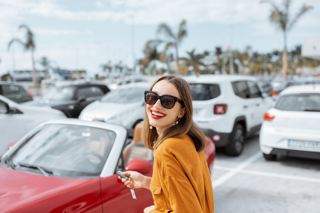 Lächelnde Frau mit Sonnenbrille vor einem roten Auto auf einem Parkplatz